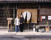  トレードマークの丸く染め抜いたのれんが掛かる「ひのきと草木染織工房」の前で加納さんとスタッフの上田久美子さん